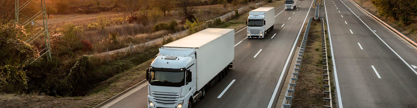 four large white lorries travelling down a main road in single file