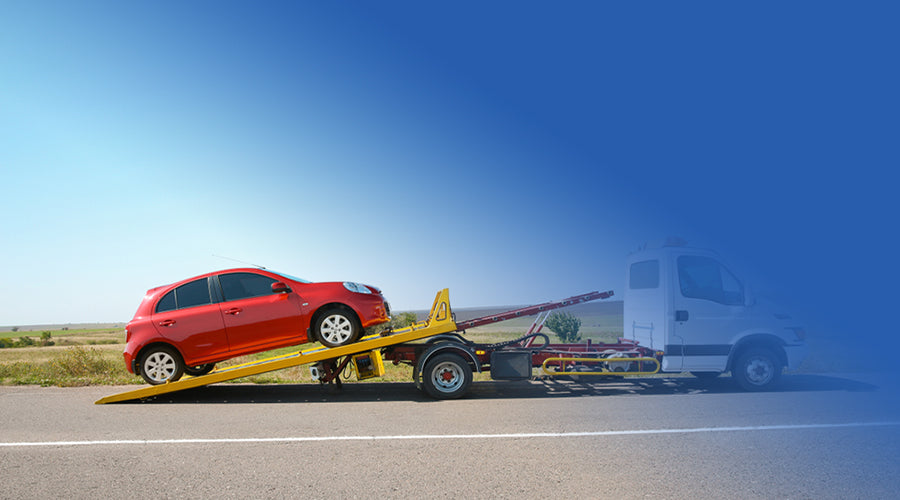 a small red car being towed along a main road by a white recovery truck using taut straps